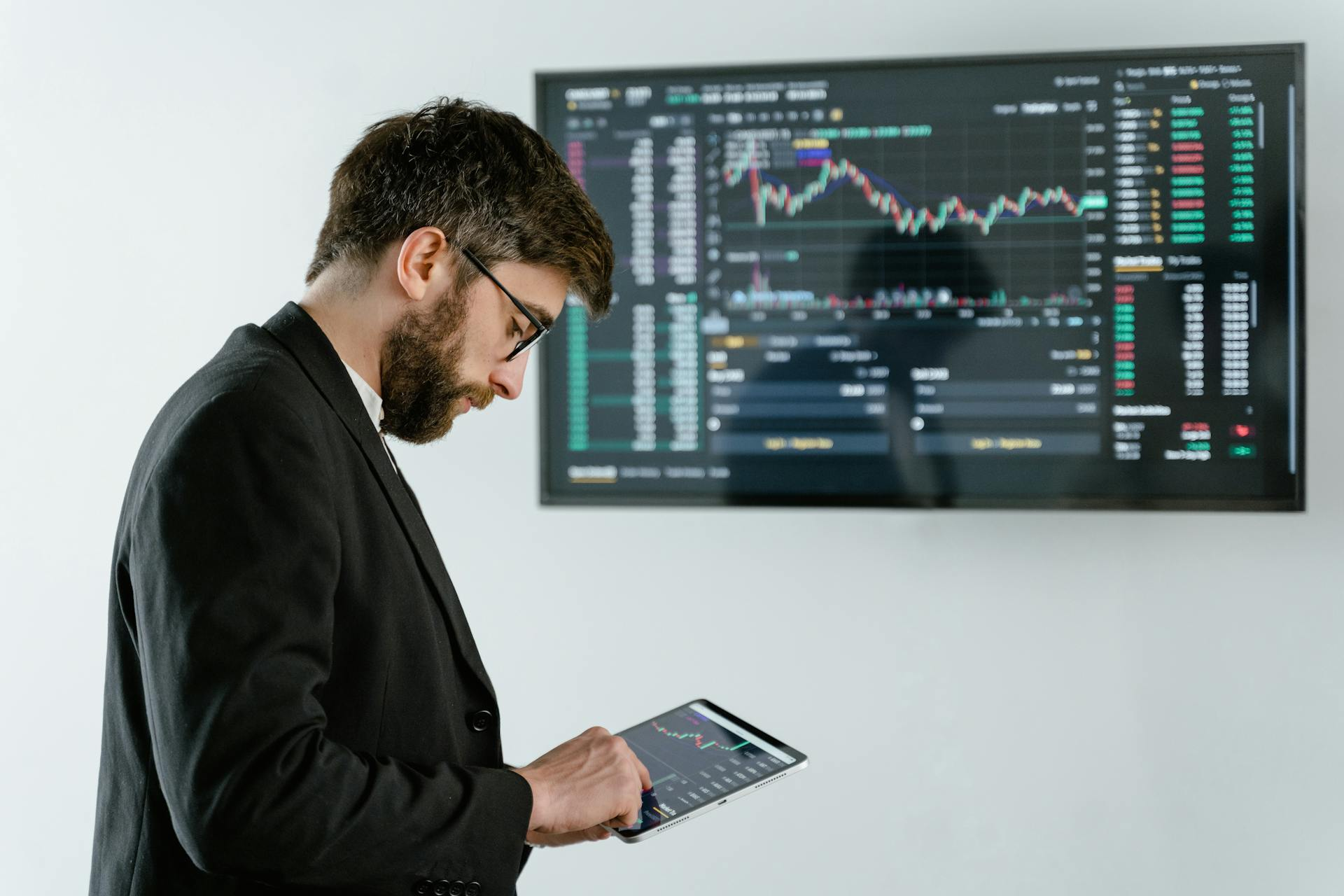 Ein Mann in einem Anzug steht vor einer Wand mit einem großen Bildschirm. Auf dem Bildschirm sind Diagramme und Daten zu sehen, die typischerweise mit Börsenkursen oder Finanzmärkten in Verbindung stehen. Der Mann hält ein Tablet in den Händen. Das Bild steht im Zusammenhang der Digital Signage Software