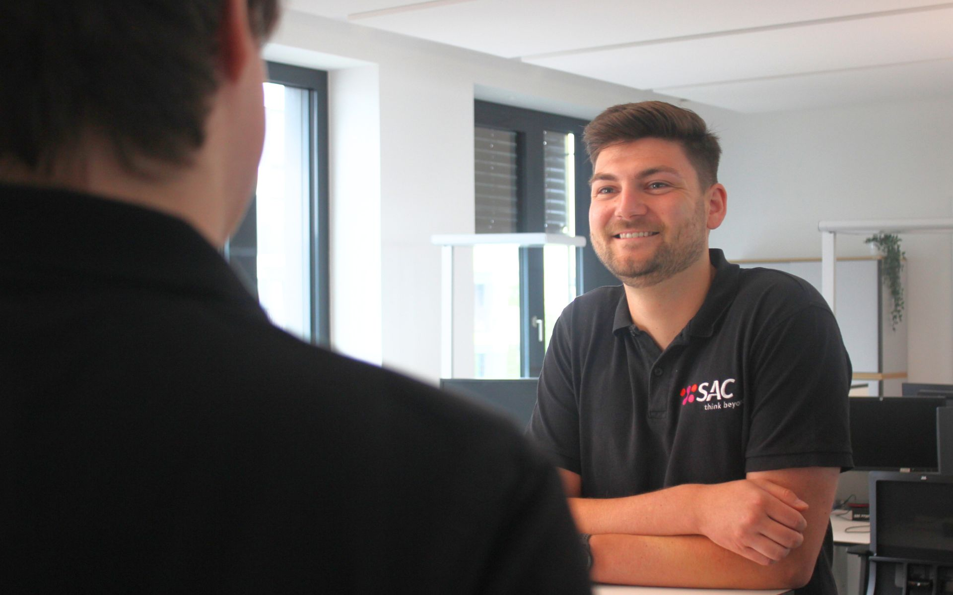 Lächelnder Mitarbeiter des IT-Service Provider SAC im Büro in einem schwarzen SAC Polohemd spricht zu einer anderen Person